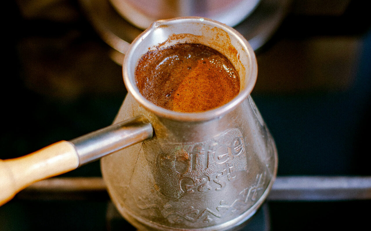 Gros plan d'une cafetière traditionnelle en métal utilisée pour préparer du café turc, avec le café en train de bouillir à l'intérieur, créant une mousse épaisse sur le dessus.