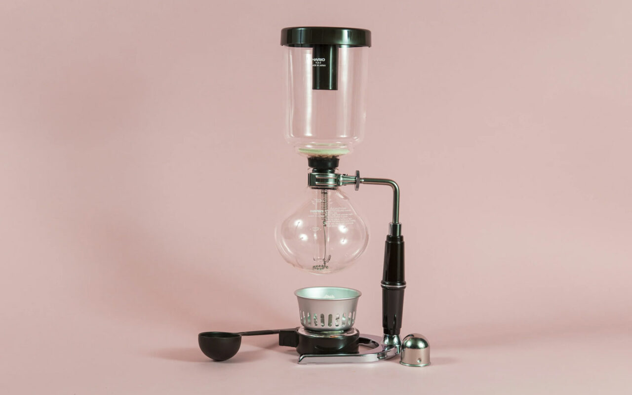 Une cafetière à dépression, également connue sous le nom de siphon, est placée sur un fond rose pâle. L'appareil est composé de deux globes en verre connectés par un tube, avec une base en métal noir et un support pour la flamme en dessous.