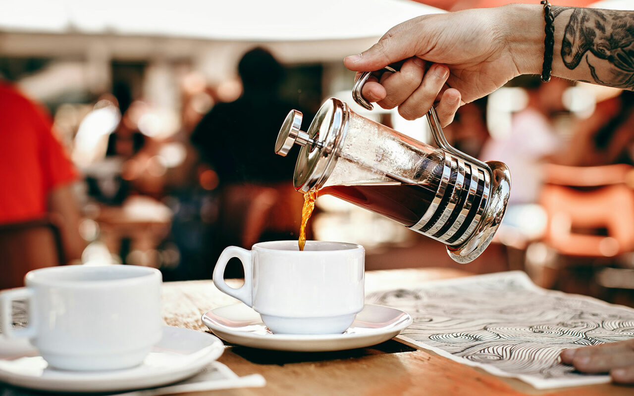 Une main tatouée verse du café fraîchement préparé d'une cafetière à piston (French Press) dans une tasse blanche, avec une deuxième tasse blanche posée sur la table à côté. En arrière-plan, on peut apercevoir une terrasse de café avec des personnes floues en train de profiter de la journée.