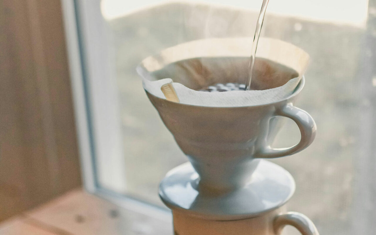 Un filtre en papier placé dans un porte-filtre en céramique, avec de l'eau chaude versée sur du café moulu pour une extraction de café en méthode V60, posée près d'une fenêtre lumineuse.