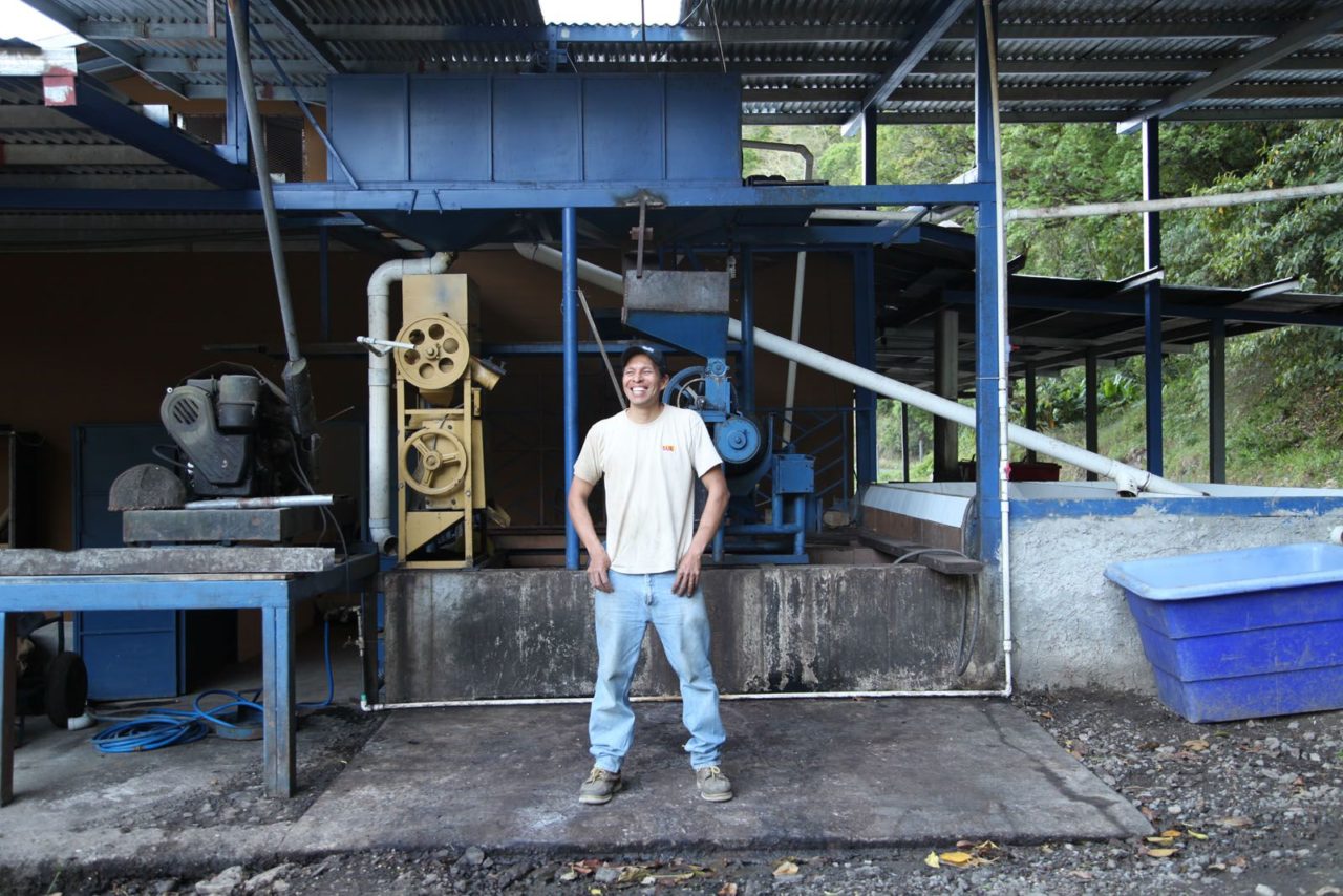 Homme souriant debout devant une machine agricole dans une structure de transformation de café en plein air.