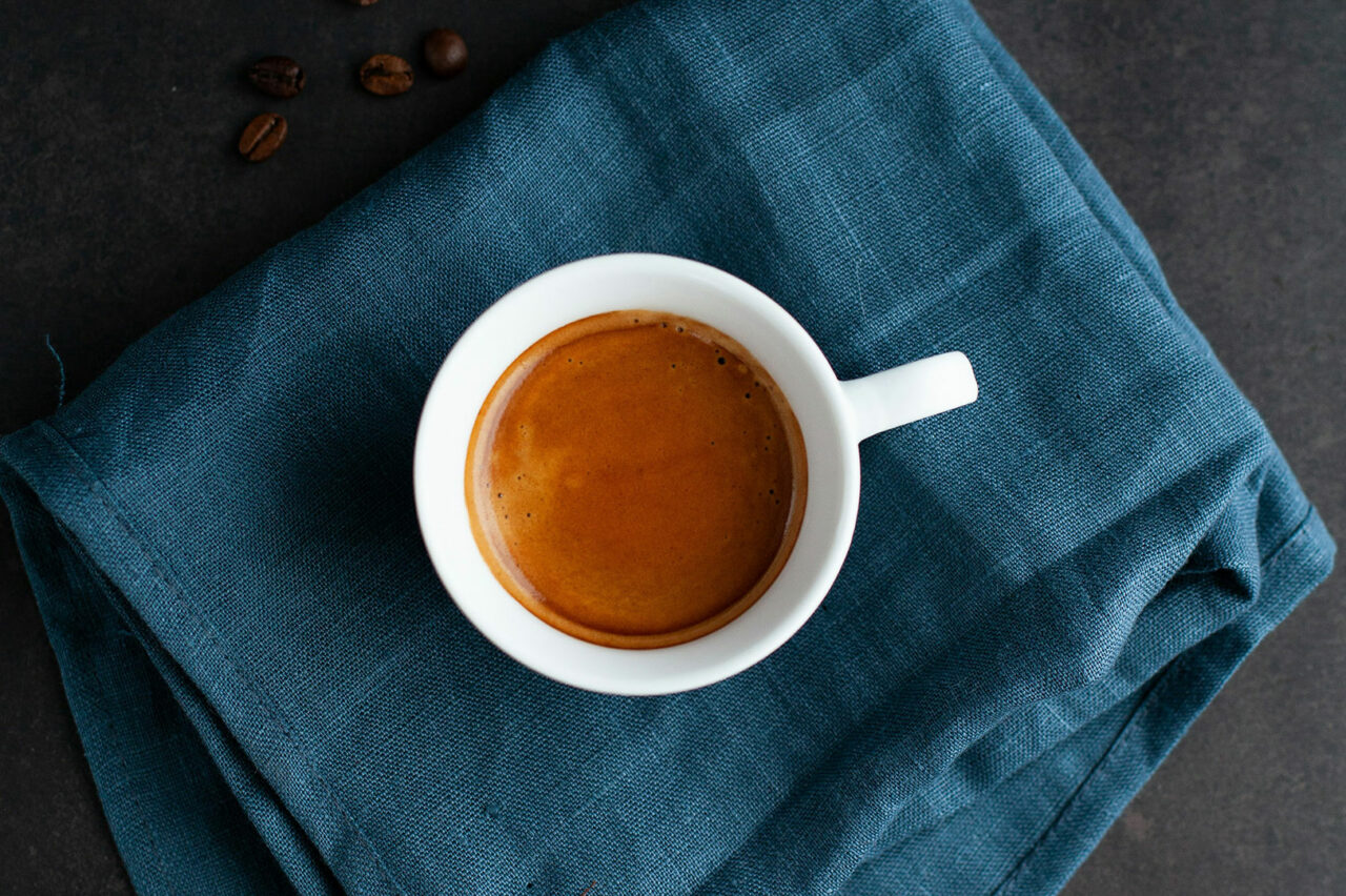 Une tasse de café espresso crémeux vue de dessus, posée sur un tissu bleu foncé avec quelques grains de café éparpillés en arrière-plan.