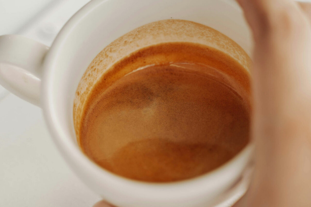 Gros plan d'une tasse blanche contenant un café espresso, montrant des traces de mousse le long des parois de la tasse. Une main tient la tasse par la poignée.