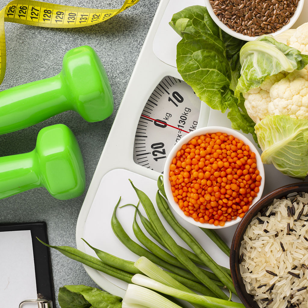 Une vue en plongée d'éléments liés à la santé et au fitness, comprenant des haltères verts, un mètre ruban jaune, une balance, et des aliments sains comme du chou-fleur, des lentilles rouges, du riz, des haricots verts et des graines de lin, disposés sur une surface grise.