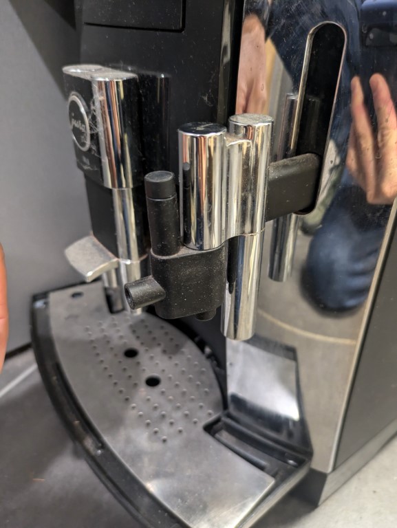 La buse à lait est correctement installée sur la machine à café JURA. On peut voir le bec verseur chromé de la machine avec la buse en place, prête à être utilisée. La machine présente quelques traces de poussière sur les surfaces métalliques.